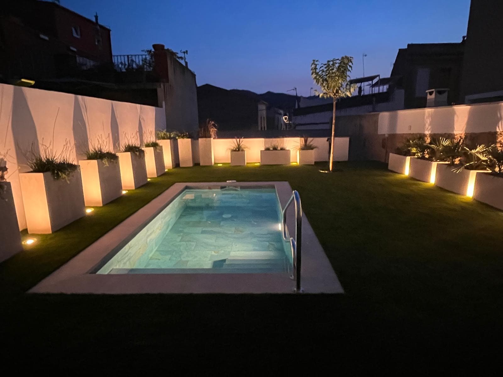 Piscina rectangular iluminada de noche, con jardineras arquitectónicas en un jardín contemporáneo